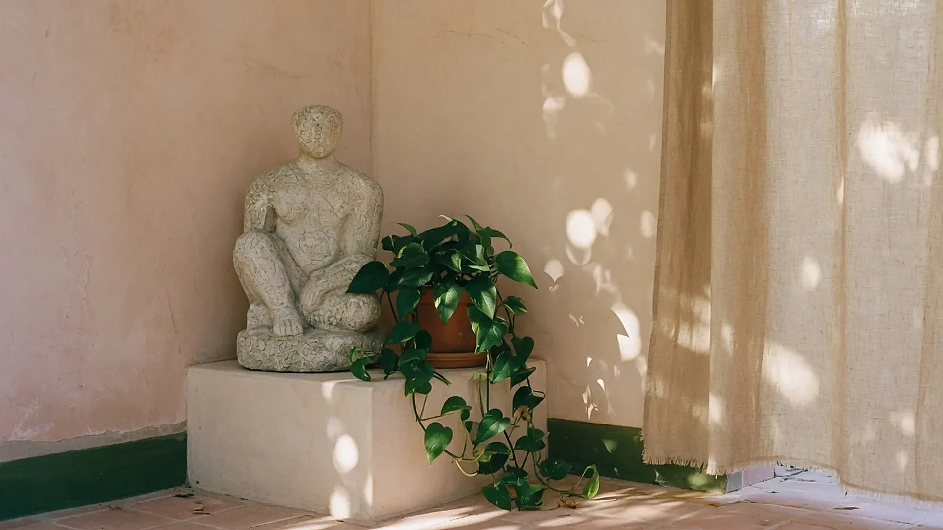 A quiet studio corner with a stone sculpture, a trailing plant and a linen curtain in afternoon sun