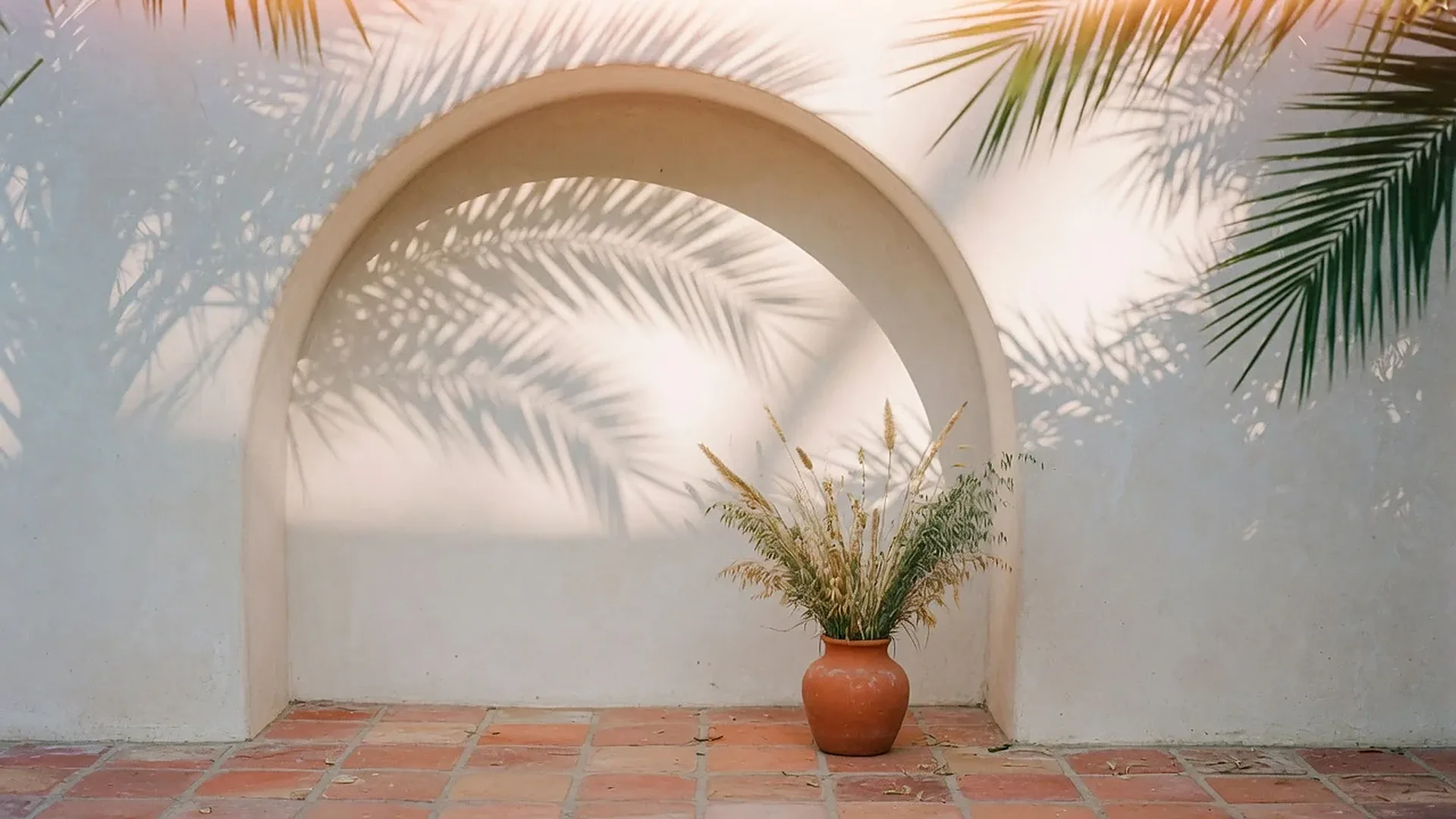 A plaster archway with soft palm-leaf shadows and a clay vase of dried grasses